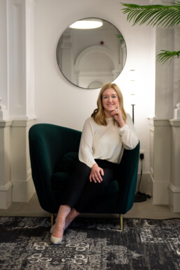A blonde white woman sat on a green velvet chair in front of a round mirror. There is a grey rug on the floor. The woman is wearing a white top and black trousers with cream pointed high heels.